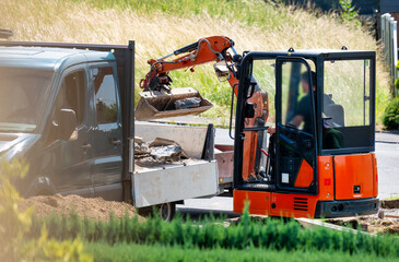 A mini excavator loads rubble into a flatbed truck during landscaping or construction work