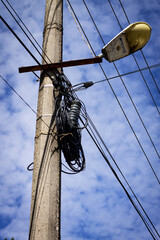 A weathered utility pole stands against a bright blue sky, entangled with a chaotic mess of black...