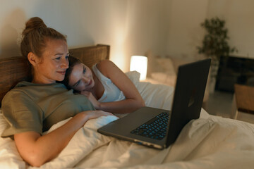 Fototapeta premium Middle age woman and daughter relaxing together in bed with laptop, enjoying wellbeing, healthy lifestyle, and warm family moments at home in cozy lighting.