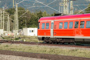 Bright red train passing through a railway yard in daylight amidst green foliage and electrical...