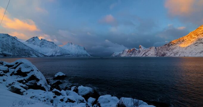Fischerboot im Fjord auf Senja in Norwegen im Winter zwischen goldener und blauer Stunde, schneebedeckte Berggipfel von warmem Licht beleuchtet, ruhige arktische Stimmung