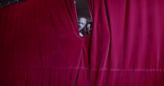 Two actors peek through the stage curtains, curious about the audience. The anticipation and excitement before a performance.