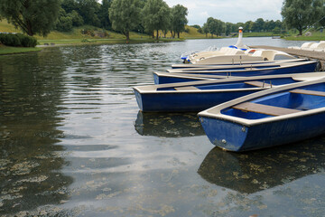 Multiple blue boats lined up by the calm waters of a lake surrounded by green trees during a sunny...