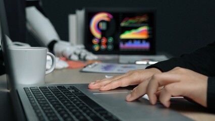 A modern workplace scene shows a person using a laptop with a coffee cup nearby, while a robotic arm interacts with vibrant data visualizations in the background.Noogenesis