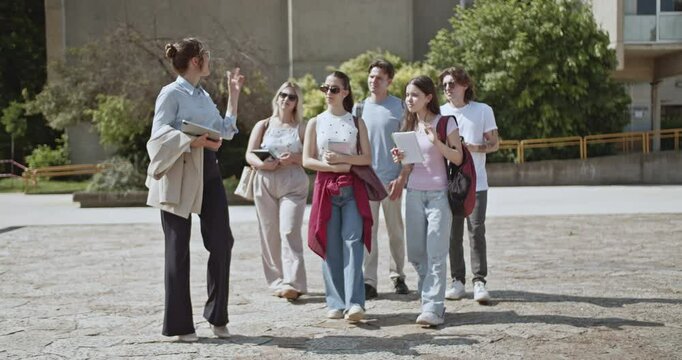 Female teacher leads a diverse group of young university students on a guided tour across the campus grounds, explaining and sharing educational insights. They are learning and engaging in discussion.