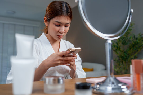 Woman white robe is focused her smartphone while sitting vanity table with skincare products. She embodies serene moment of self care after refreshing shower, emphasizing importance of skin