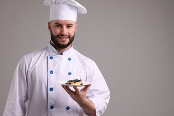 Happy confectioner in uniform holding delicious tart with blueberries on light grey background