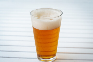 A full pint of golden beer with thick foam is placed on a white slatted surface, creating a refreshing summer drink moment. There is space for text on the side.