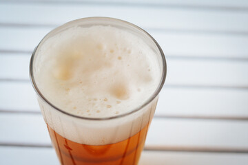 A full pint of golden beer with thick foam is placed on a white slatted surface, creating a refreshing summer drink moment. There is space for text on the side.