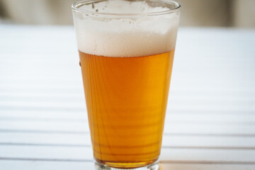 A full pint of golden beer with thick foam is placed on a white slatted surface, creating a...