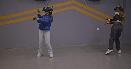 Two young women friends are having fun playing games in virtual reality. They are wearing VR headsets and holding controllers.