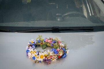 A colorful wreath of wildflowers rests on the wet hood of a car, reflecting the light of a recent...