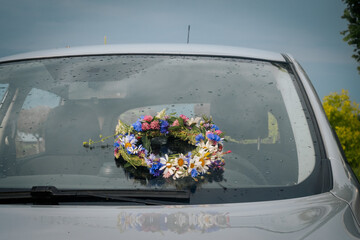 A colorful wreath of wildflowers rests on the wet hood of a car, reflecting the light of a recent...