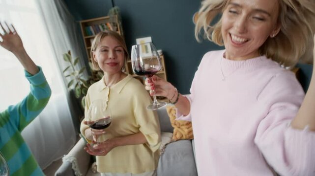 Slow motion dutch angle of three middle-aged female friends dancing and clinking wine glasses in living room