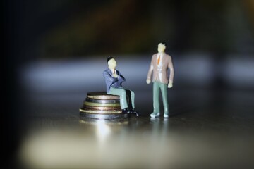 duas miniaturas plasticas de homen vestindo roupas sociais como executivos, em espaco vazio,  um deles seuntado sobre pilha de moedas, piso de madeira com foco bem direcionado e profundidade de campo.