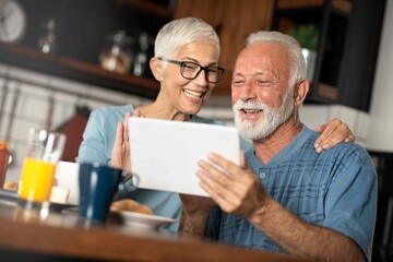 Happy older mature couple having breakfast using digital tablet in kitchen.