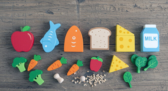 A colorful assortment of wooden toy food items including an apple fish bread cheese milk broccoli carrots spinach and beans arranged on a rustic wooden surface
