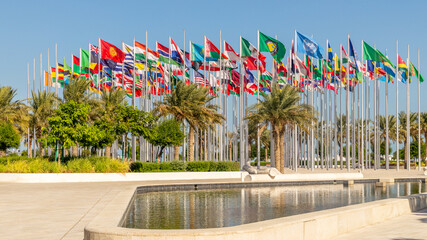 Many world's countries national flags waving on the wind, with UN, EU, GCC, Belarusian, Kyrgyz, Mexican banners with palms and pond in the foreground, Doha, Qatar