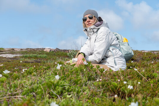 Woman in a gray beanie, white jacket, and sunglasses sitting among arctic vegetation in Teriberka Nature Reserve under a clear blue sky