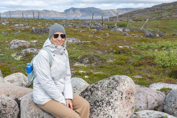 Smiling woman in a gray beanie, white jacket, and sunglasses sitting on a large granite rock in the...