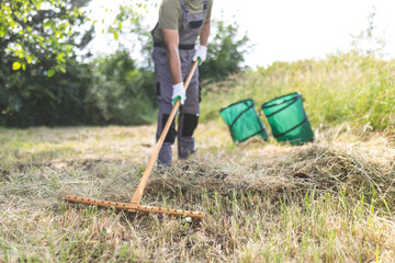 Person raking grass in a sunny outdoor field near green waste bins during a late spring afternoon