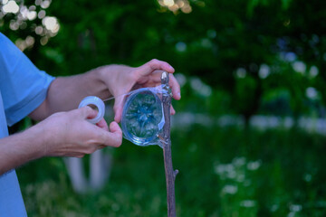 Man uses transparent adhesive tape to attach a cut plastic bottle to a wooden stick in a grassy outdoor environment, likely for a DIY or garden-related purpose.