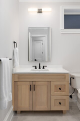 A bathroom detail with a white oak cabinet, white countertop with a black faucet, light mounted above the mirror, and brown tile flooring.
