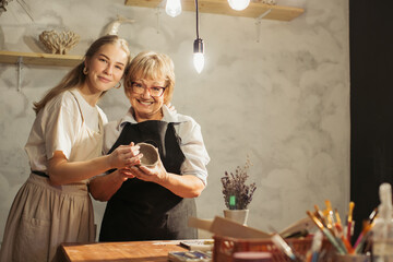 Senior woman and young apprentice holding clay vase in pottery workshop
