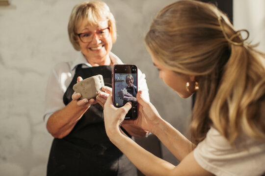 Potter showing handmade clay vase while student is taking photo with smartphone