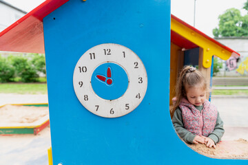 playground clock on colorful blue playhouse with smiling girl playing in sandbox outdoor...