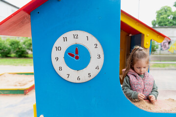 playground clock on colorful blue playhouse with smiling girl playing in sandbox outdoor...