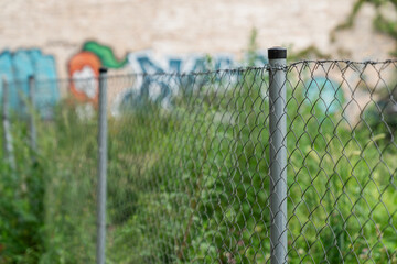 urban chain link fence with overgrown grass and graffiti wall background, city boundary barrier in...