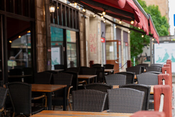 empty outdoor cafe terrace with wicker chairs and wooden tables under red canopy awning in city street, peaceful european restaurant patio scene with no people, urban dining area atmosphere