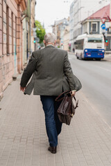 senior businessman walking on city sidewalk with leather briefcase wearing suit jacket and blue...