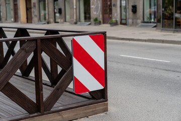 urban traffic warning sign with red and white diagonal stripes on wooden barrier near sidewalk,...
