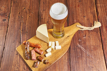 A glass of ice-cold golden beer on a rustic wooden board, accompanied by a cheese and beer sausage appetizer.
