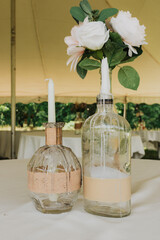 Two upcycled glass bottle centerpieces with dripping wax taper candle and white rose–peony floral...