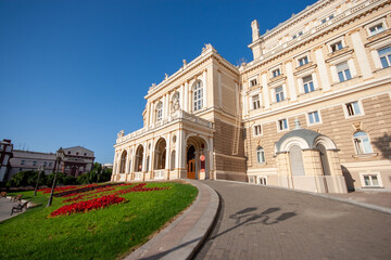 Fototapeta premium Grand Opera and Ballet Theatre building with classical architecture, surrounded by vibrant red flowers and a lush green lawn under a clear blue sky, showcasing culture and heritage