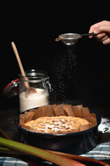 Hand sprinkles powdered sugar over a baked rhubarb cake in its tin, surrounded by rustic kitchen props in a dark moody setting.
