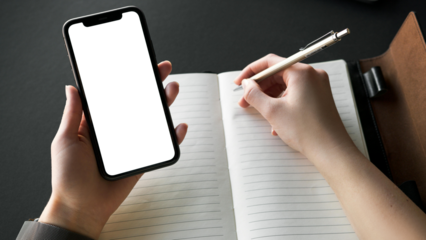 Hands hold a phone and write in a notebook on a desk, capturing modern productivity and multitasking capabilities for business, education, or lifestyle.