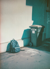 Old, worn backpack thrown away next to a garbage container. Back to school, forgotten knowledge conceptual background.