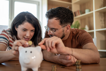 Young couple saving money together, inserting coins in piggy bank at home