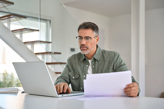 Serious busy middle aged mature man wearing eyeglasses holding paper bill checking insurance invoice or tax document calculating expenses working online at home using laptop computer.