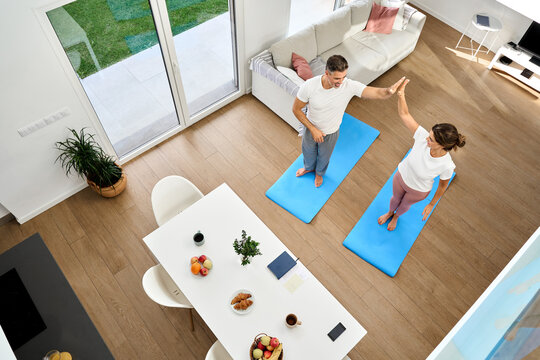 Fit healthy mature man and woman standing on mats doing morning training exercises in modern house living room. Happy middle aged couple giving high five after workout at home. Top view from above