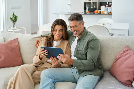 Candid photo of happy middle aged couple using digital tablet relaxing on couch at home. Smiling mature man and woman holding tab browsing internet on pad device sitting on sofa in living room.