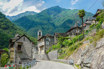 Winding road leads through a traditional stone village in an alpine valley, flanked by old houses,...