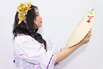 Naklejka premium A young Hispanic woman wearing a traditional costume is looking at a sombrero while holding it in her hands, against a white background. Colombian culture concept
