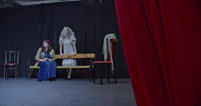 Two young actresses wait backstage on a bench. One looks through binoculars while the other looks towards the stage. Red curtain partially visible.
