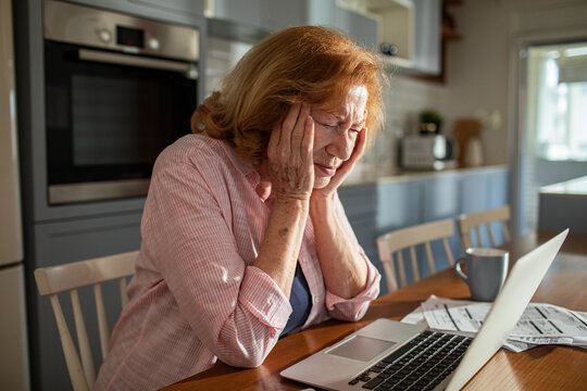 Senior woman stressed while looking at laptop and bills at kitchen table