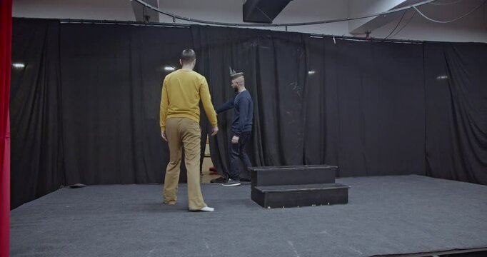Two actors conclude their rehearsal on a stage. They exit through a gap in the backdrop curtains. The stage is dark and empty, ready for the main performance.